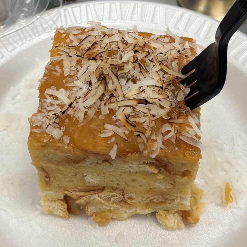 A slice of Italian dessert with a golden top and toasted coconut flakes, on a white plate with a black fork.