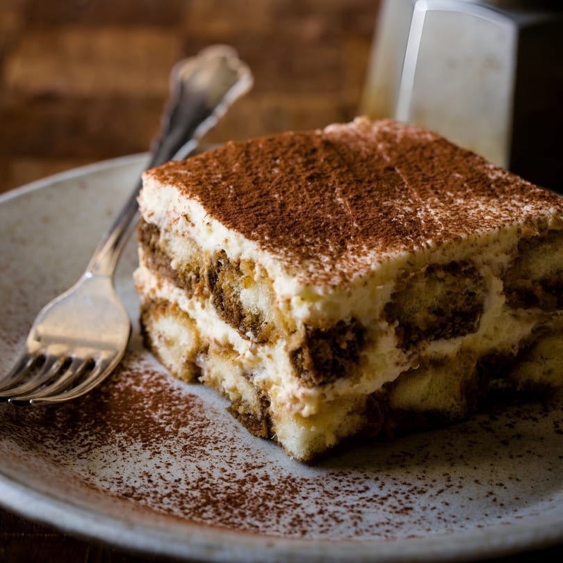 A slice of tiramisu with cocoa powder on top, served on a white plate with a fork.