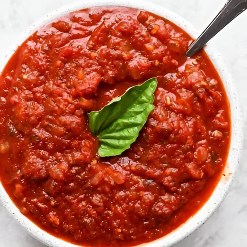 Close-up of a bowl of chunky red marinara sauce with a fresh basil leaf and a spoon.