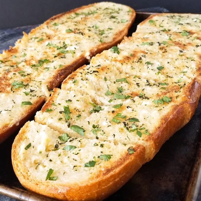 Two golden-brown garlic bread loaves, topped with melted cheese and fresh parsley, on a dark baking sheet.
